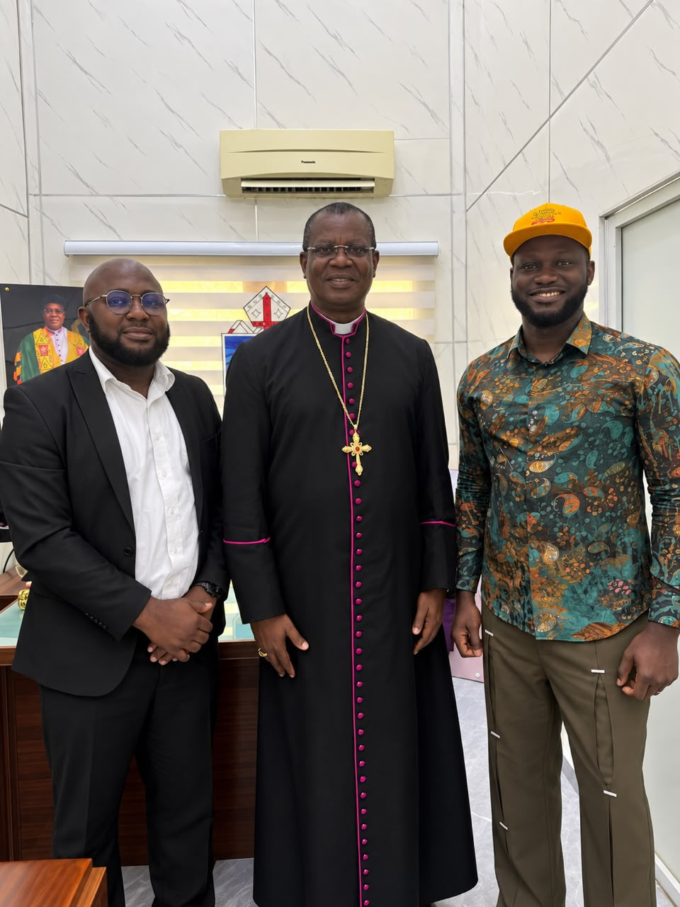 Three men standing in an office; the man in the centre wears a bishop's cassock and pectoral cross.