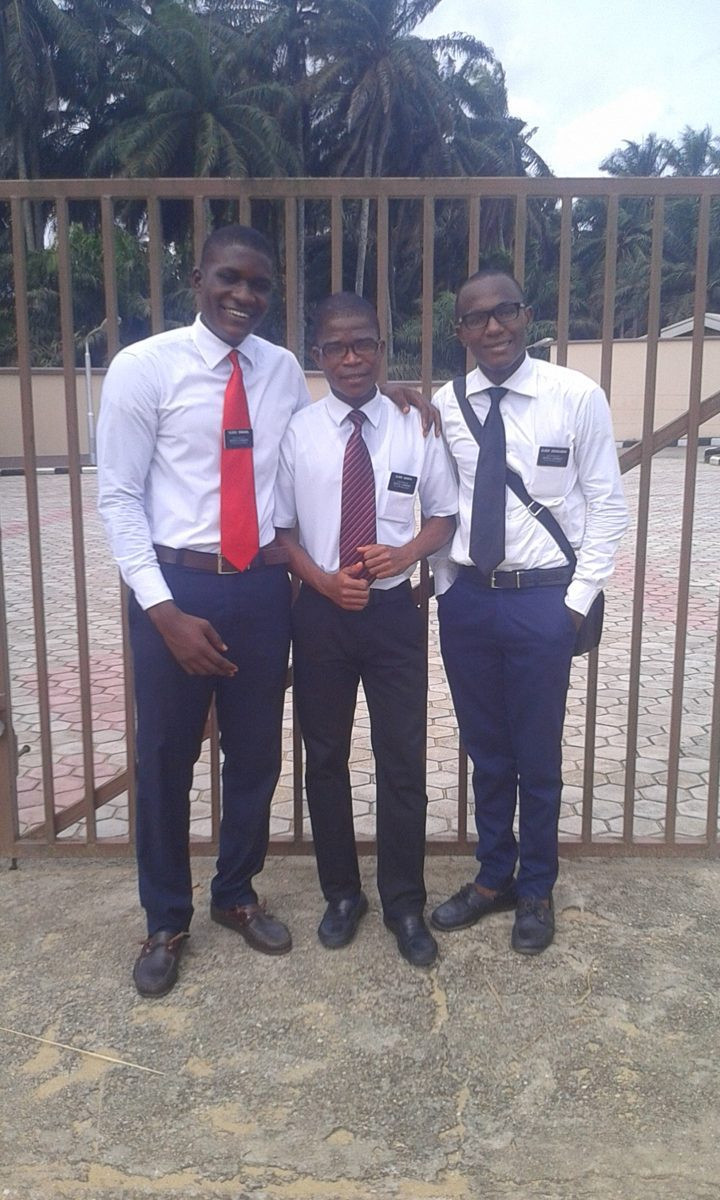 Three young men in white shirts and ties standing in front of a fence.